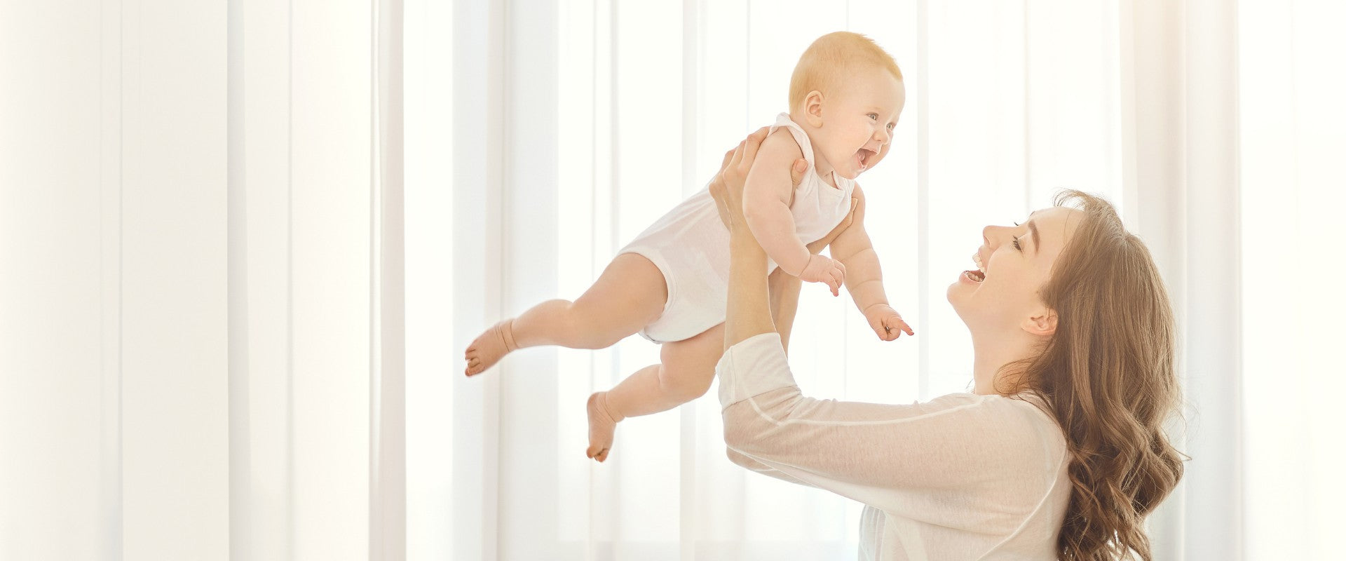 Smiling mother lifting her baby in the air, both dressed in soft, neutral-toned outfits from a Babywonders gift set — capturing a joyful bonding moment.