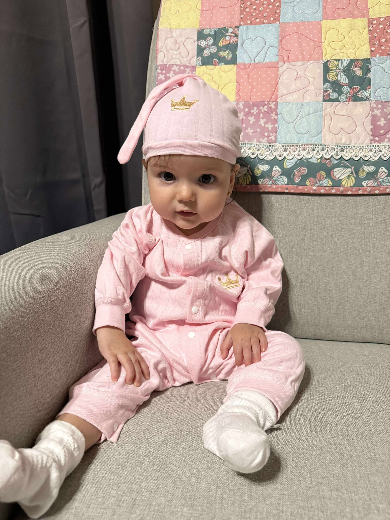 Baby girl wearing pink BabyWonders gift set and hat with gold crown emblem, sitting on a sofa
