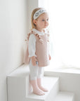 Baby girl in soft pink outfit and ribbed socks standing by window in natural light