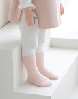 Baby girl in soft pink outfit and ribbed socks standing by window in natural light