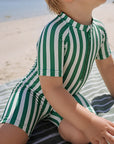 Baby wearing green stripe short sleeve zip swim romper sitting on beach towel, lightweight breathable summer baby swimwear