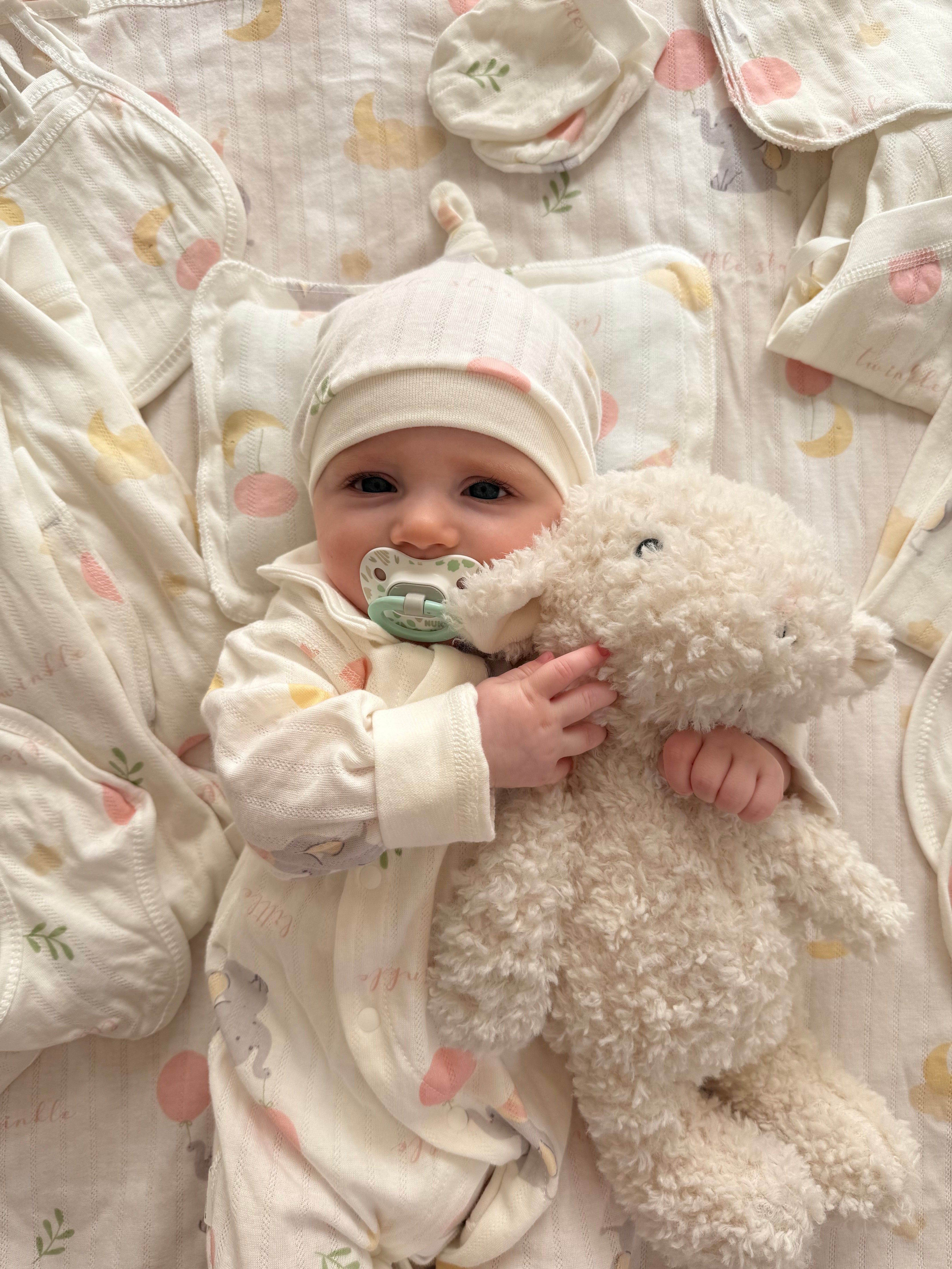 Baby lying on BabyWonders essentials Gift set in soft neutrals, cuddling Lumi the lamb plush toy and wearing a printed onesie with dummy