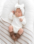 Baby wearing classic white Peter Pan collar romper with brown velvet bow baby socks lying on soft neutral baby lounger bedding
