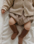 Baby wearing brown ribbed knee-high cotton socks with neutral outfit on bed