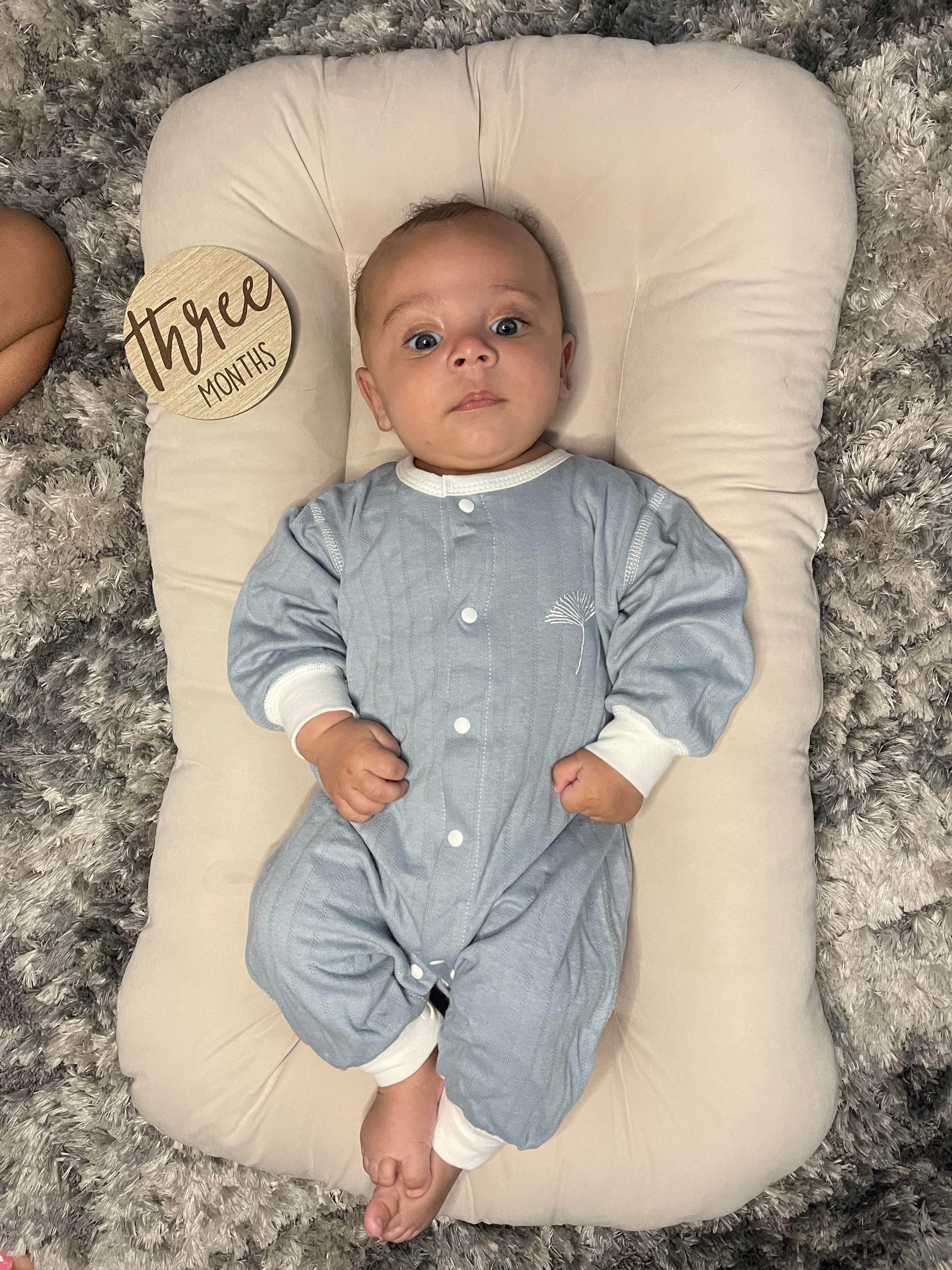 Baby in a blue onesie lying on a cushion with a 'Three Months' milestone card. Babywonders gift set from cozy night 28pieces. Gift Idea for newborn baby