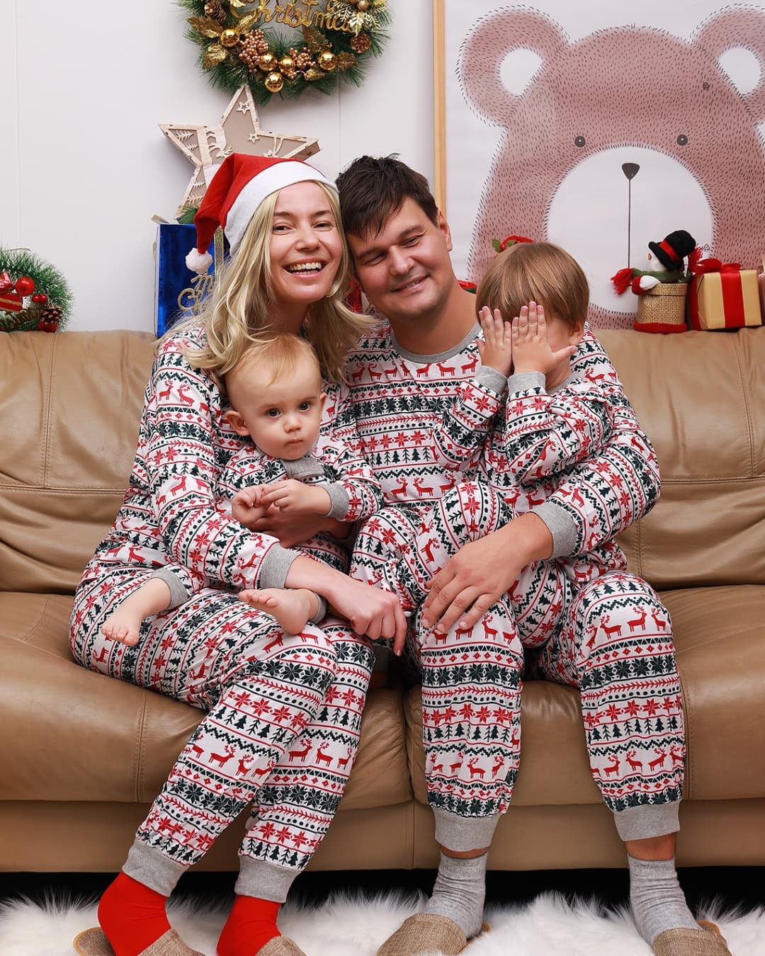 Family wearing matching red and white Christmas pajamas with reindeer and snowflake pattern, sitting on a sofa during holiday celebration.