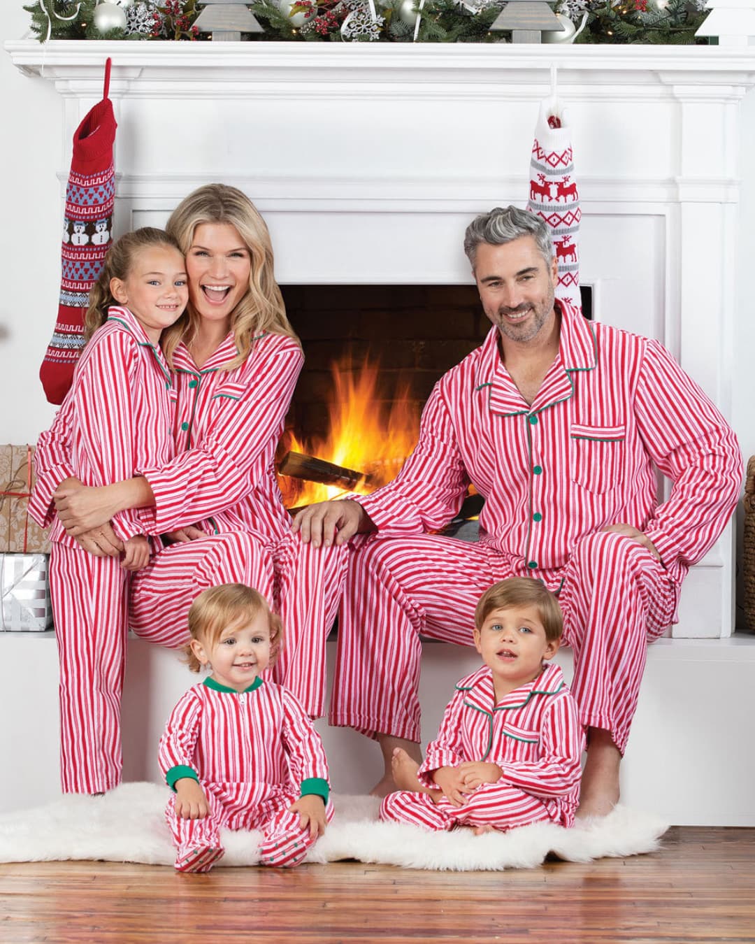 Family wearing matching red striped Christmas pajamas sitting by fireplace
