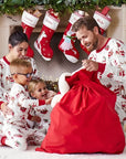 Happy family in matching Christmas pajamas with Santa print sitting by fireplace, opening red gift bag.