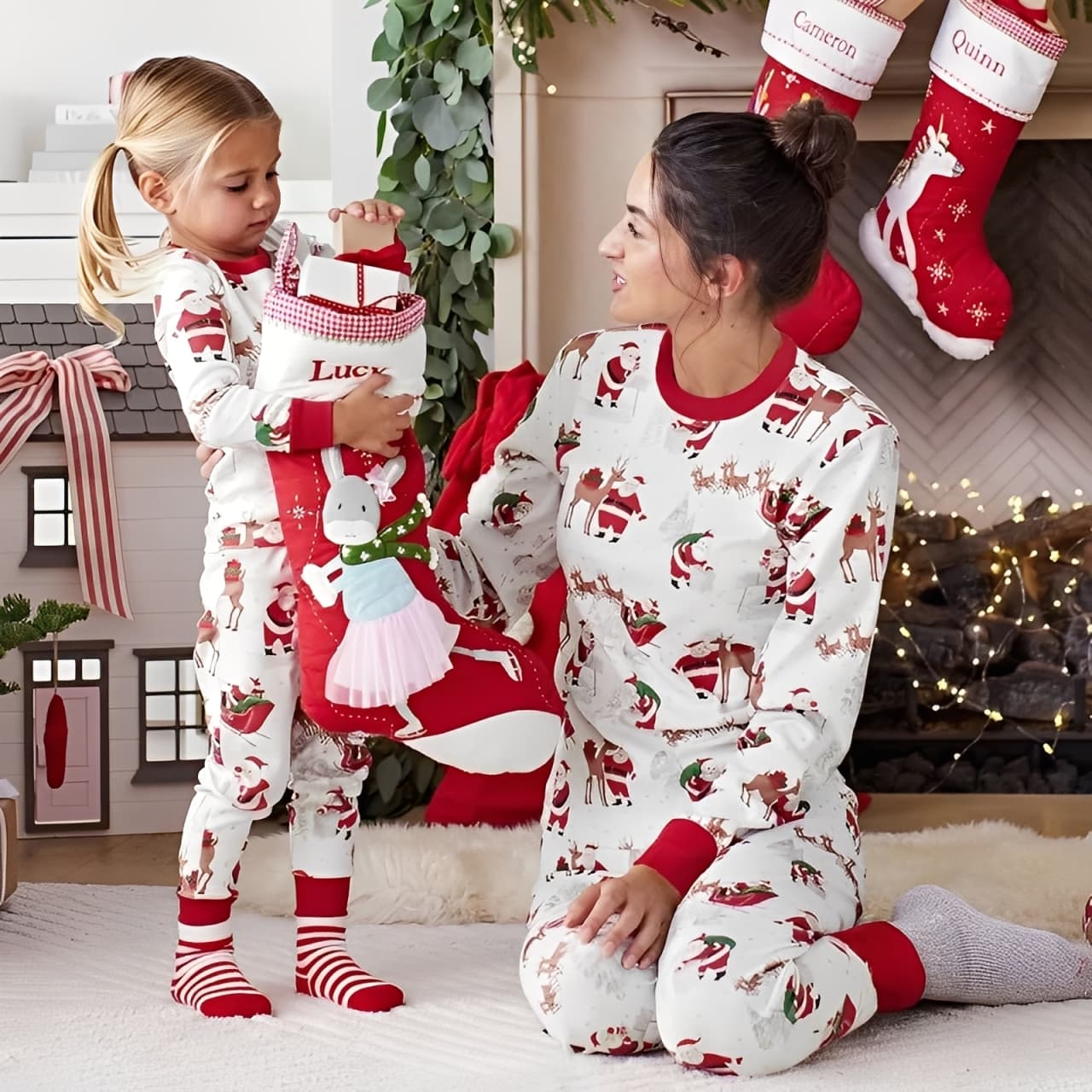 Mother and daughter wearing matching family Christmas pajamas with Santa and reindeer print, opening stocking by fireplace.