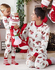 Mother and daughter wearing matching family Christmas pajamas with Santa and reindeer print, opening stocking by fireplace.