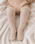 Baby wearing cream ribbed long lace knee-high socks on cosy neutral bedding