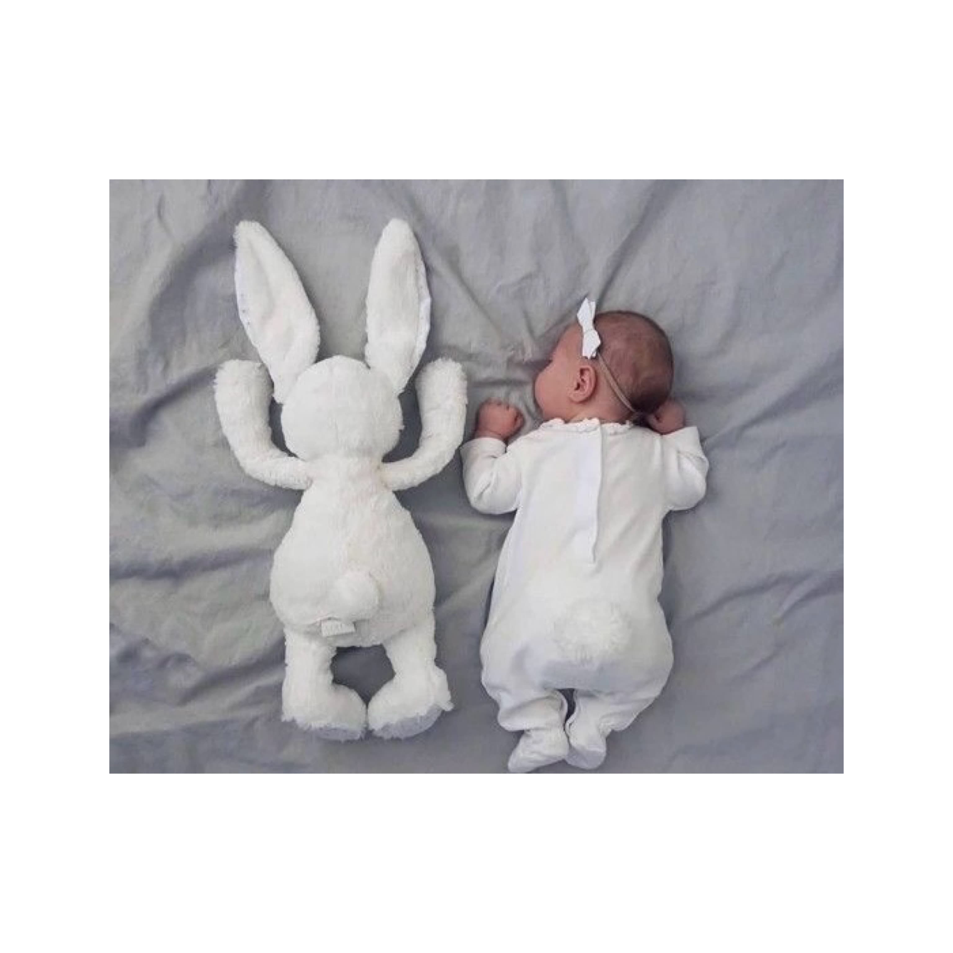 Newborn baby lying on a bed in a white onesie with a headband, placed next to a large white stuffed bunny in a similar position.