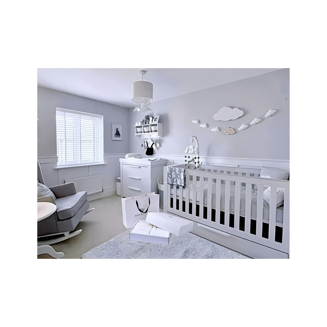 A serene, modern nursery decorated in soft grey and white tones, featuring a crib, rocking chair, changing unit, and cloud-themed wall decor. A BabyWondersUK gift box set is placed on the rug, suggesting a thoughtful and stylish baby gift for the space.