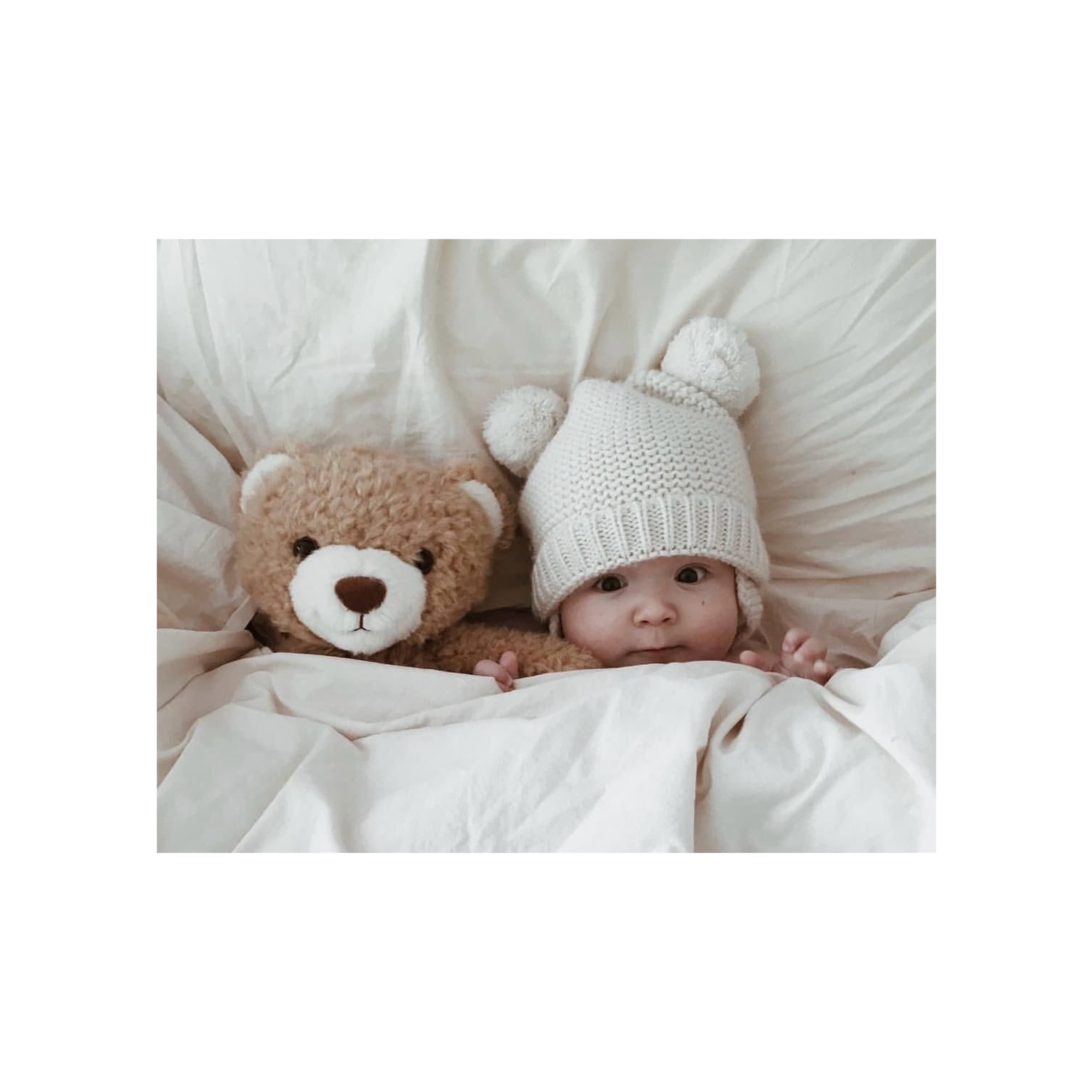 Baby lying in bed wearing a knitted hat with bear ears, snuggled under a blanket beside a teddy bear.