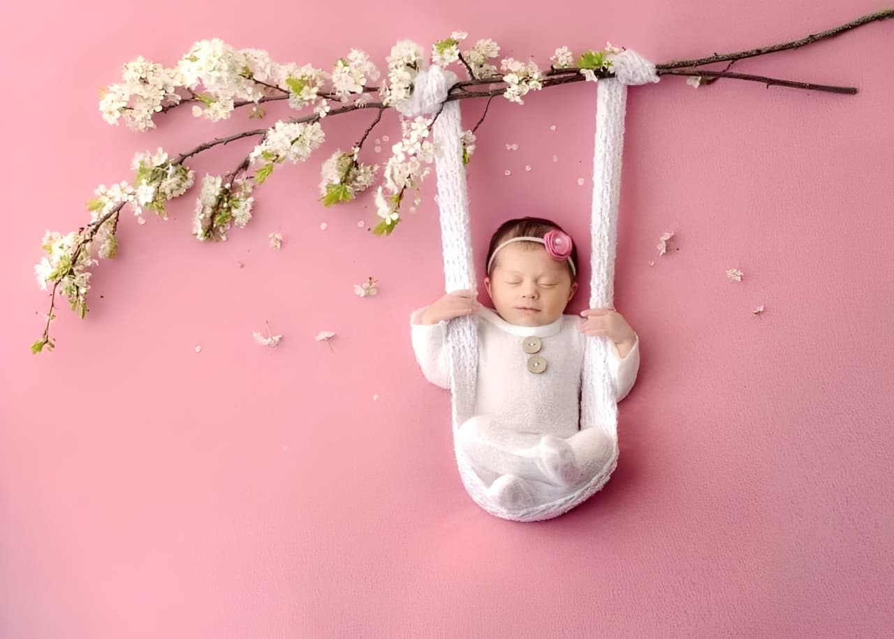 Newborn baby girl in pink outfit lying on floral swing, safe and comfortable baby clothes gift idea.