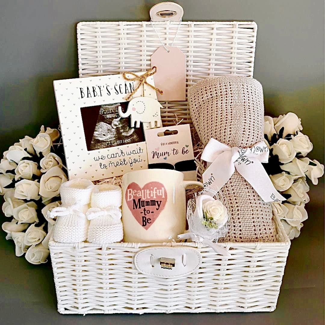 A beautifully arranged white wicker baby gift hamper featuring a ‘Mummy to Be’ mug, ultrasound photo frame, knitted baby booties, soft blanket, faux roses, and a tag, ideal for baby showers and expectant mothers.