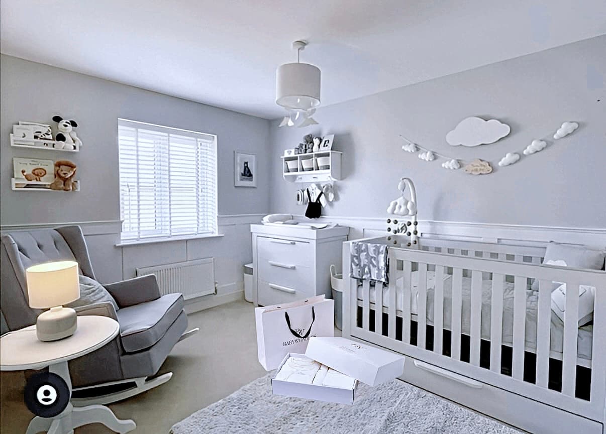 A serene, modern nursery decorated in soft grey and white tones, featuring a crib, rocking chair, changing unit, and cloud-themed wall decor. A BabyWondersUK gift box set is placed on the rug, suggesting a thoughtful and stylish baby gift for the space.
