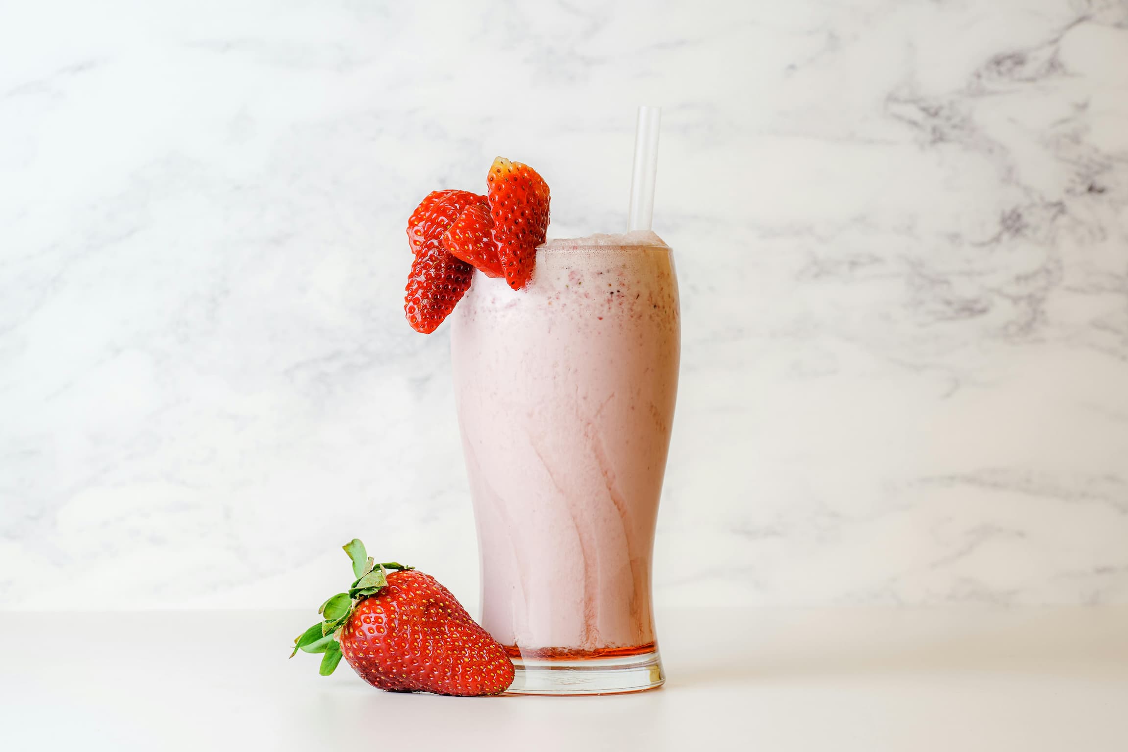 A creamy pink strawberry smoothie served in a tall glass, garnished with fresh strawberries on the rim and placed on a white marble surface — a refreshing and nutritious drink ideal for postpartum recovery.