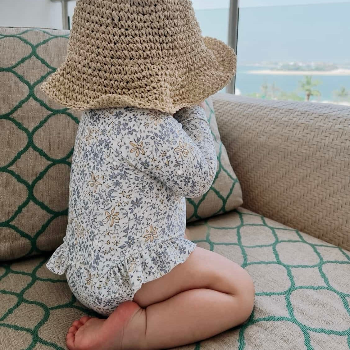 Baby wearing beige floral long sleeve swimsuit sitting indoors with straw hat, soft breathable toddler swimwear
