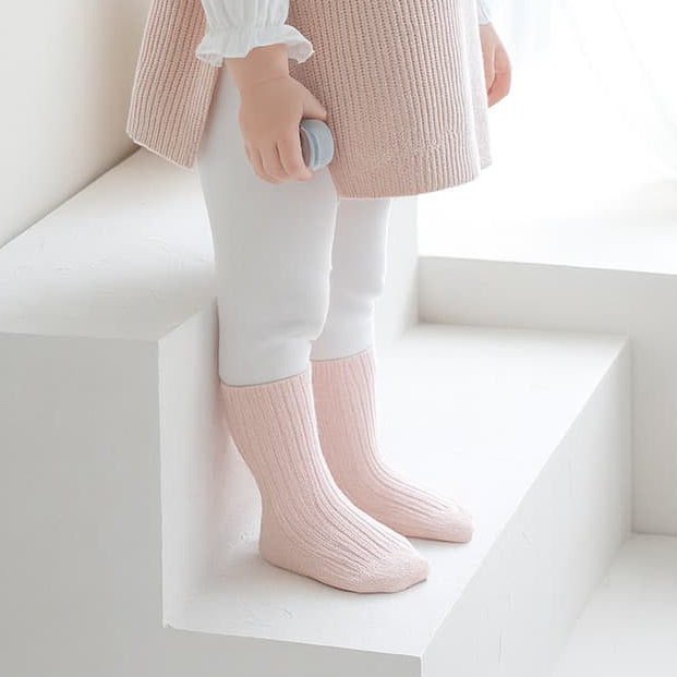 Baby girl in soft pink outfit and ribbed socks standing by window in natural light
