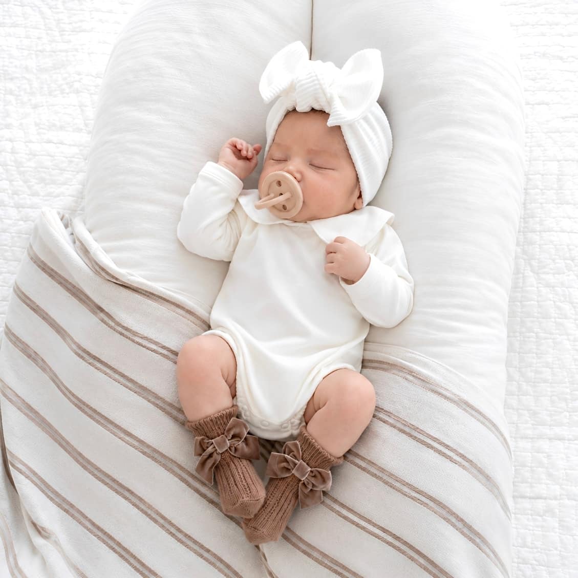 Baby wearing classic white Peter Pan collar romper with brown velvet bow baby socks lying on soft neutral baby lounger bedding