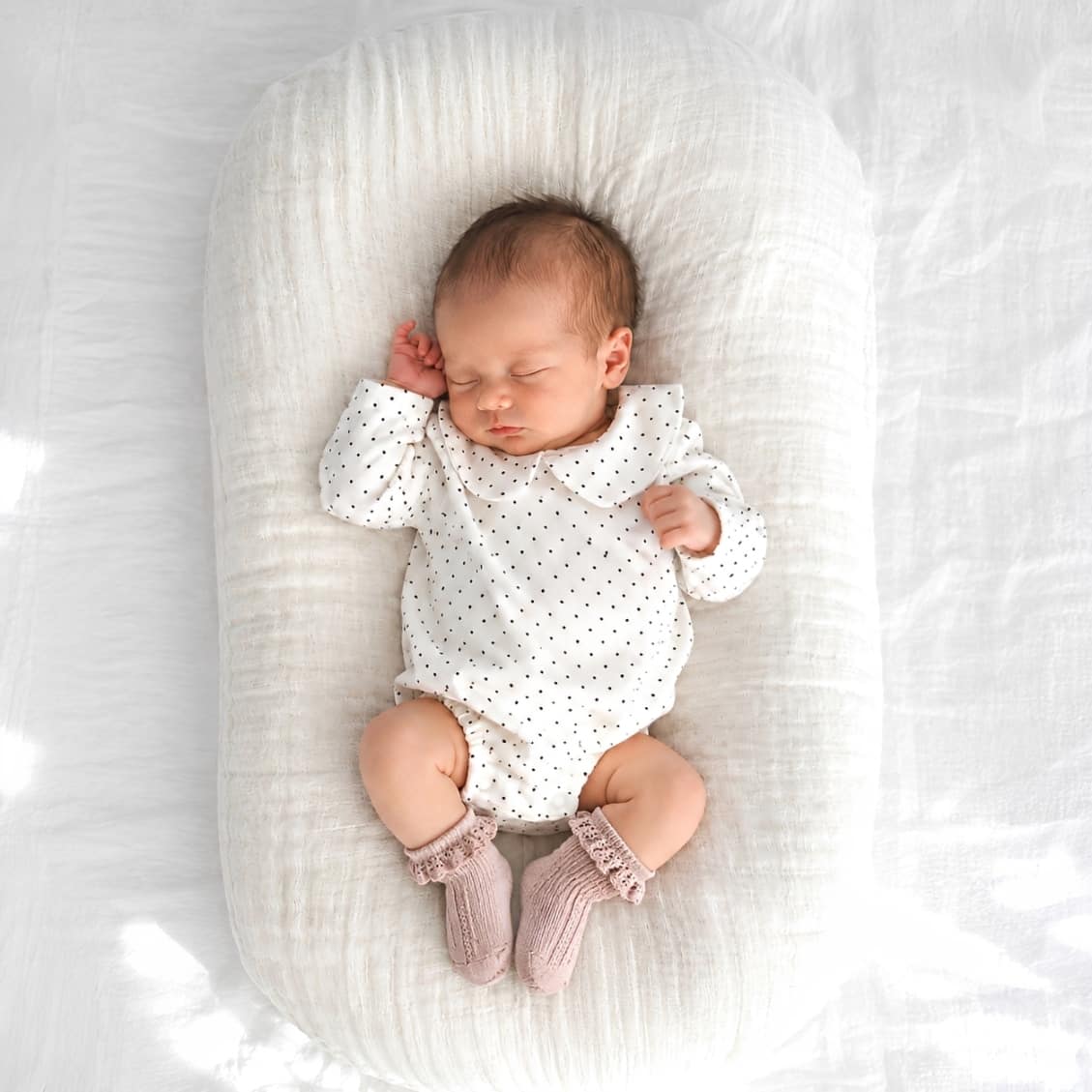 Baby wearing white polka dot Peter Pan collar romper with blush pink frill lace baby socks lying on soft neutral baby nest bedding