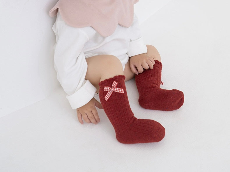 Infant sitting wearing red socks with cute checkered bow, holiday style baby outfit.
