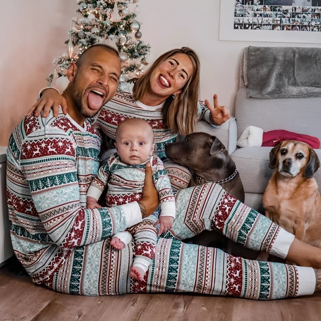 Family wearing matching Christmas pajamas with baby and dogs in front of Christmas tree
