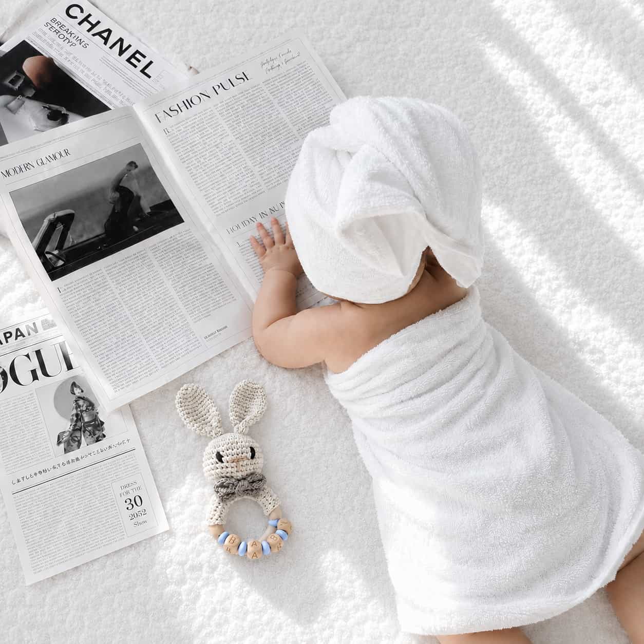 Newborn baby lying on a soft white blanket beside a crochet blue rabbit teether with personalised wooden beads, a thoughtful baby shower gift and newborn essential in the UK.