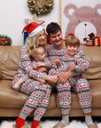 Family in coordinated holiday pajamas featuring red reindeer and festive patterns, posing together at home with Christmas decorations.
