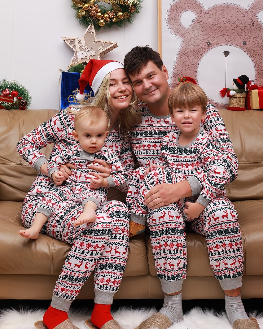 Parents and kids dressed in festive matching Christmas pajamas with red reindeer and snowflake print, smiling together on a couch.
