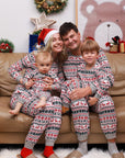 Parents and kids dressed in festive matching Christmas pajamas with red reindeer and snowflake print, smiling together on a couch.
