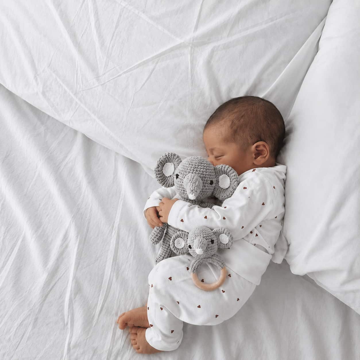Newborn baby cuddling a crochet elephant toy and wooden elephant rattle set on a soft white nursery bed.