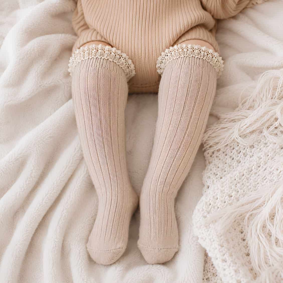 Baby wearing cream ribbed long lace knee-high socks on cosy neutral bedding