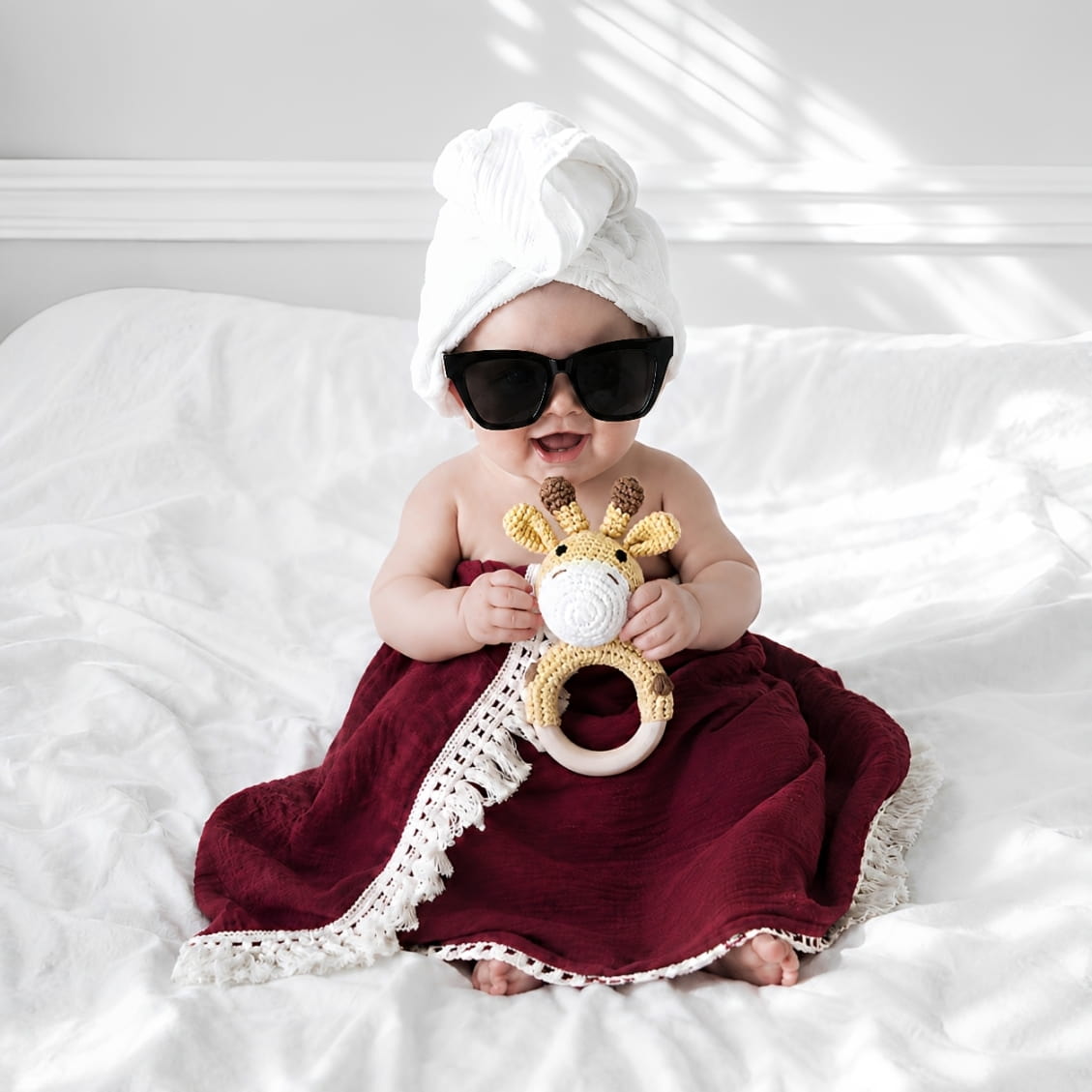 Newborn baby wrapped in a tassel burgundy baby blanket holding a crochet giraffe teether, a thoughtful baby shower gift and newborn essential in the UK.