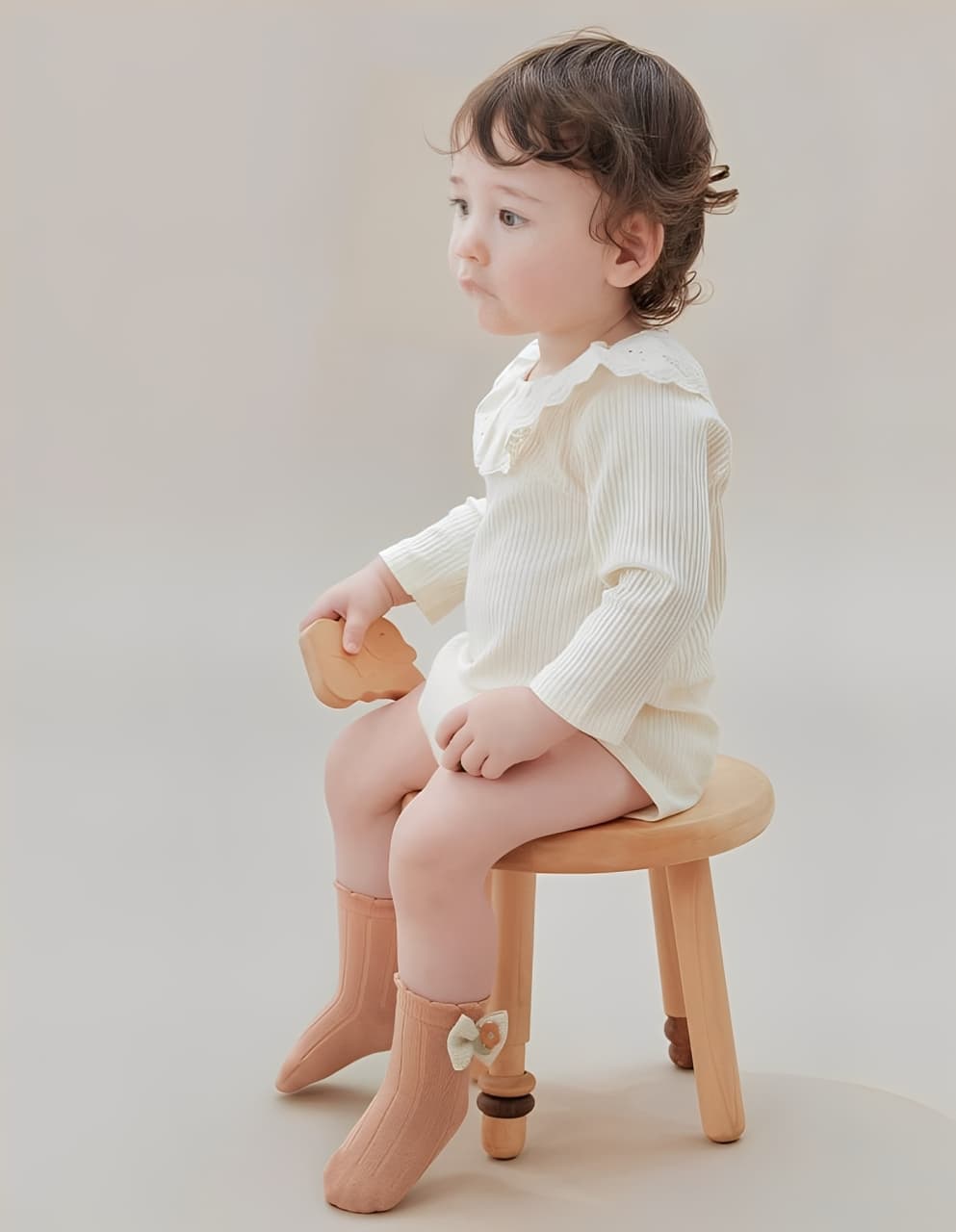 Baby wearing peach bow cotton socks, sitting on a wooden stool in neutral outfit.